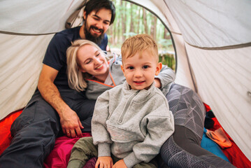 family mom dad and little boy on a hike lie and sit in a tent, hugging, laughing, having fun and...