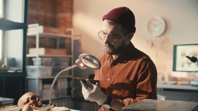Concentrated archaeologist in gloves and glasses inspecting ancient spearhead under magnifying lamp at workplace in laboratory