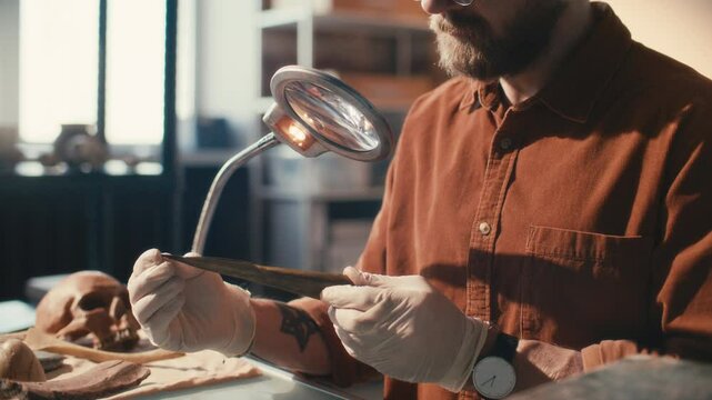 Archaeologist in gloves observing spearhead under magnifying lamp as researching historical artifacts in laboratory. Midsection, close-up shot
