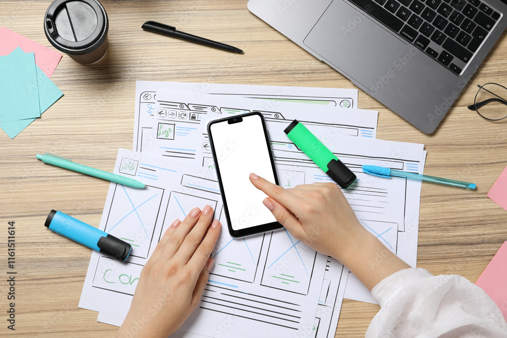Sticker website designer using smartphone at wooden table with wireframes and laptop, top view