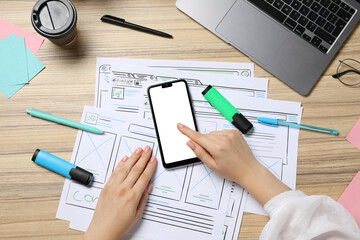 Website designer using smartphone at wooden table with wireframes and laptop, top view