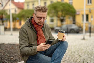 Young Attractive Man On Park Bench Drinking Coffee And Using Smartphone