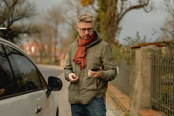 Obraz premium Young Attractive Man With A Car On A Street Checks An Incoming Message On Smartphone