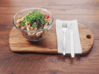 Fresh salad with tuna, tomato and dill in a glass round bowl standing on a wooden board and table. Easy healthy portion of food.