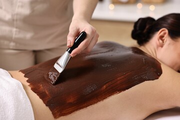 Chocolate body wrap. Spa worker applying mask onto woman's back in salon, closeup