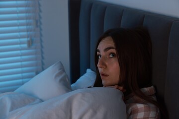 Fear of darkness. Scared young woman lying on bed in room at night, space for text