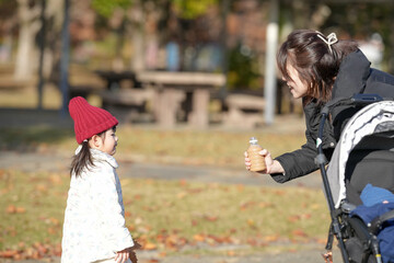 A 3-year-old Japanese girl and a woman in their 30s, wearing down jackets, drink warm milk tea from plastic bottles on a cold winter afternoon in a park in Hakata City, Fukuoka Prefecture.