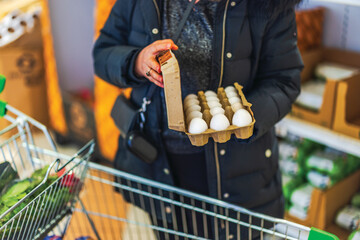 Woman in black winter coat holding carton of white eggs while shopping in grocery store aisle....