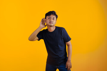 young asian man is eavesdropping with his ears. yellow background