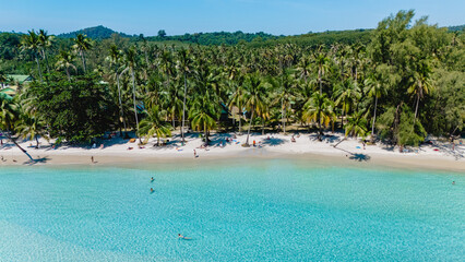 Crystal turquoise waters gently lap against the soft white sand shore in Koh Kood, Thailand. Sunbathers relax under the shade of palm trees, enjoying a tranquil tropical day on the beach.