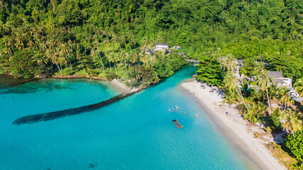 Crystal-clear waters embrace the shores of Koh Kood, Thailand, as sun-kissed beaches meet verdant hills. Visitors enjoy tranquil moments in this tropical paradise, surrounded by nature's beauty.