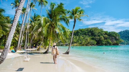 Warm sun illuminates the soft sands as a woman walks along the shoreline of Koh Kood. Lush palm...