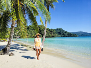 A woman strolls leisurely on the soft sandy beach of Koh Kood, surrounded by swaying palm trees and crystal-clear waters. The vibrant scenery captures a serene tropical paradise.