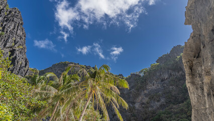 Blue sky with clouds are framed by mountains. Cliff tops and steep furrowed slopes. Green tropical...