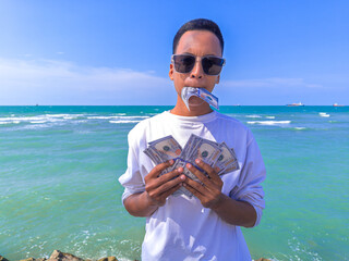 Asian guy with white shirt and wear glasses. Concept Confident man holding a 100 dollar bill in his hand, man hold 100 USD. Summer sea water background. focus on man. noisy, exposure, similar others