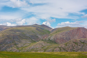 Beautiful layered landscape with multicolor hills and mountains in sunlight under clouds in blue sky. Scenic alpine view to sunlit colorful mountain range of many vivid colors in changeable weather.