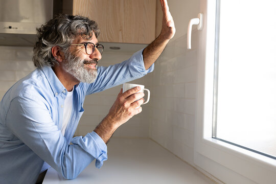 Happy mature man relaxing at home having morning coffee looking out the kitchen window waving to friends. Copy space