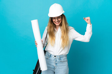 Young architect woman with helmet and holding blueprints isolated on blue background doing strong...
