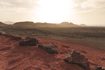 Fototapeta premium Vast volcanic landscape in Lanzarote with red sand, lava rocks, and mountains under a hazy sky at Timanfaya National Park