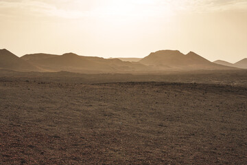 Vast volcanic landscape in Lanzarote with red sand, lava rocks, and mountains under a hazy sky at ...