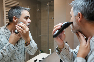 Mature man trimming beard with electric razor in bathroom mirror