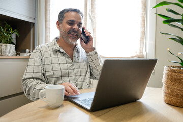 Smiling man working from home using laptop and talking on phone