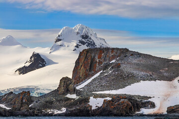 Shoreline on the Antarctic Peninsula, early morning. Rocky shore with Penguins; behind snow covered mountains; clouds, blue sky above. 
