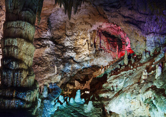 A cave with stalagmites and stalactites, illuminated with colorful lighting that highlights the natural rock formations. The textured walls and mineral structures create an ethereal atmosphere. 