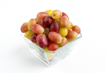 Fresh red and yellow grapes in a glass container, placed on a minimalist white background.

