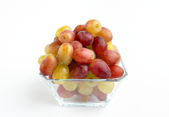 Fresh red and yellow grapes in a glass container, placed on a minimalist white background.
