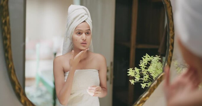 Indian upset young adult gen z woman lady wrapped towel on head and body look mirror feel sad facial pores skin dry cheek teen girl female worried for acne pimple touch face heath care at indoor home