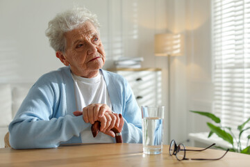 Thoughtful senior woman feeling lonely at table with glass of water indoors