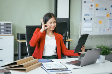 An Asian businesswoman works at her desk in a modern office, smiling while using a tablet to review financial analysis reports, manage accounting tasks, and oversee banking and loan documentation