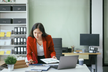 An Asian businesswoman works at her desk in a modern office, smiling while using a tablet to review financial analysis reports, manage accounting tasks, and oversee banking and loan documentation