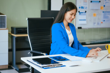 An Asian businesswoman works at her desk in a modern office, smiling while using a tablet to review...