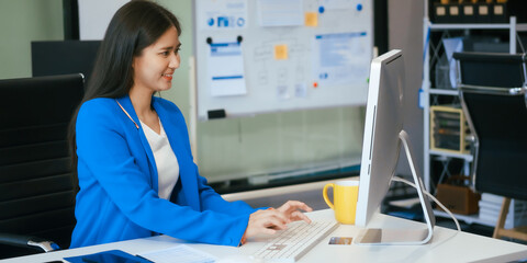 An Asian businesswoman works at her desk in a modern office, smiling while using a tablet to review...