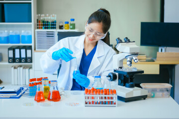 A young female scientist, wearing a lab coat,safety glasses, gloves, examines a test tube...