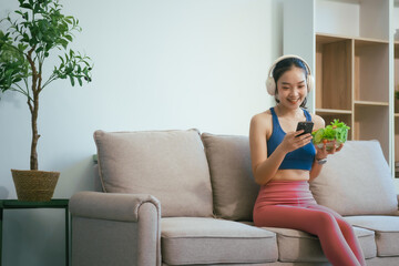 A happy Asian woman in sportswear sits on a sofa at a table, enjoying a salad bowl, fruits, fresh vegetables, orange juice, and energy supplements after a home fitness workout