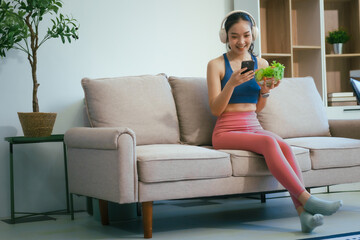 Obraz premium A happy Asian woman in sportswear sits on a sofa at a table, enjoying a salad bowl, fruits, fresh vegetables, orange juice, and energy supplements after a home fitness workout