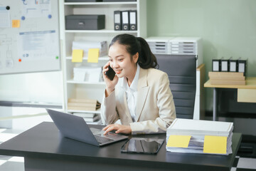 An Asian businesswoman sits at her office desk, smiling happily while working on her laptop. She reviews documents, plans new business strategies, and manages tasks efficiently to achieve success