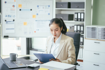 An Asian businesswoman sits at her office desk, smiling happily while working on her laptop. She reviews documents, plans new business strategies, and manages tasks efficiently to achieve success