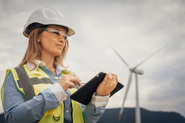 An experienced female engineer is inspecting a wind turbine site, using her tablet to assess the equipments functionality and ensure compliance with strict sustainability standards © YURIMA