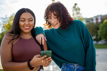 Two young women smiling while using a smartphone outdoors in a park