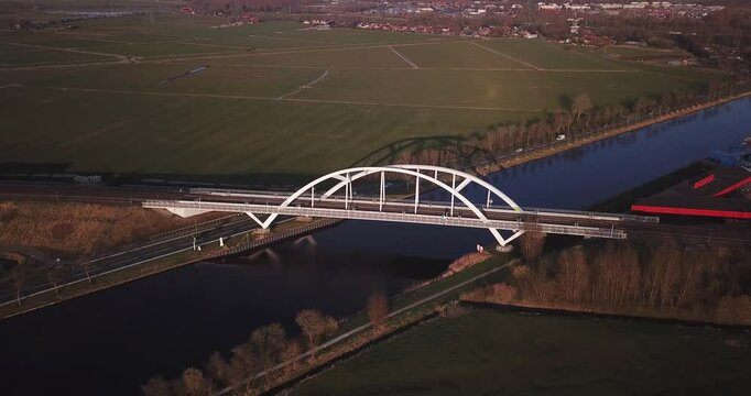 Spring evening. The camera moves towards a white arched railway bridge over a canal. At this time, a train is traveling across the bridge from left to right.