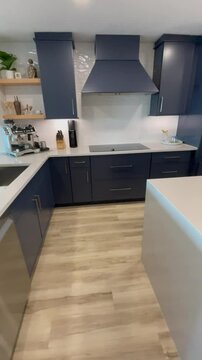 Vertical video of a modern coastal kitchen in a beachy luxury condo. The newly built custom kitchen is designed with blue cabinets, white waterfall island, white countertops, and oak wood accents.