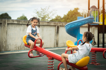 Two little girls are playing on a seesaw. One of them is wearing a white shirt and the other is...