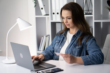 A woman is actively engaged in online shopping while using her laptop and a credit card, showcasing a modern lifestyle where technology and finance intersect in a cozy indoor workspace