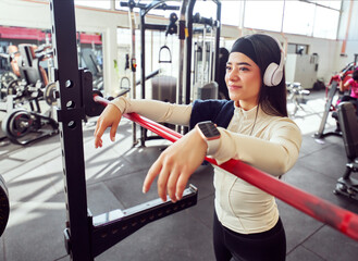 Fototapeta premium Portrait of a Latina woman in the gym with headphones next to a barbell, health and wellness