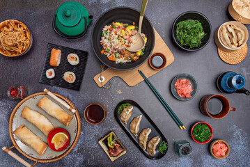 Various bowls with condiments, rice, gyozas, spring rolls and sauces on a grey stone table