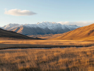 A beautiful mountain landscape featuring snow-capped peaks, expansive grasslands, and a clear blue sky with minimal clouds, illuminated by soft natural light.